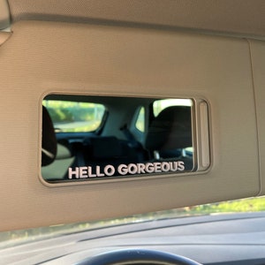 Peut inclure: Une visi&egrave;re pare-soleil de voiture avec un miroir et le texte "HELLO GORGEOUS" en lettres blanches.