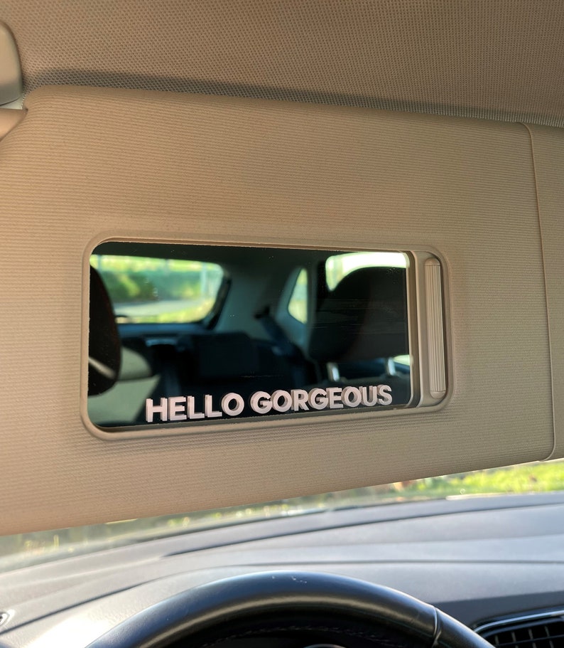 Peut inclure: Une visi&egrave;re pare-soleil de voiture avec un miroir et le texte "HELLO GORGEOUS" en lettres blanches.