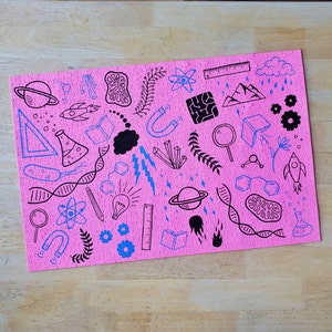 May include: Pink jigsaw puzzle with black and blue illustrations of science-related objects, including a beaker, a DNA strand, a magnifying glass, a rocket, and a light bulb. The puzzle also includes the text "Shroom Queens".