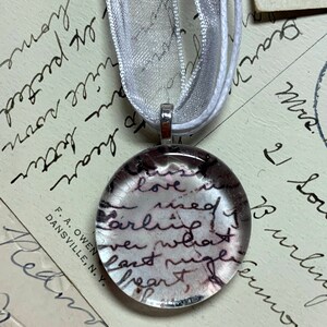 May include: A glass pendant necklace with a silver chain. The pendant is round and contains a piece of paper with handwritten text that reads "love is need early" and other words. The pendant is on a white background.