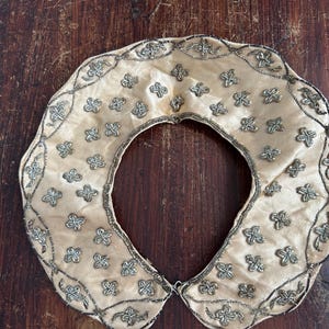 May include: Cream-colored vintage collar with intricate silver-toned floral beadwork. The collar is circular with a large opening in the center and a scalloped edge. The collar is likely made of silk or satin.