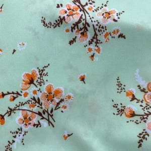 May include: Close-up of a light green fabric with a repeating floral pattern. The design features clusters of orange and white flowers with brown stems and small leaves. The overall aesthetic is delicate and botanical.