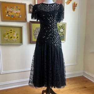 May include: A black formal dress with short sleeves and a lace overlay. The bodice is embellished with sequins, and the skirt features a tiered lace design. The dress is displayed on a mannequin.