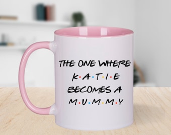 May include: White ceramic mug with a pink interior and handle. The mug features the text "THE ONE WHERE KATIE BECOMES A MUMMY" in black, with the name "KATIE" in colorful letters. A fun gift for a new parent.