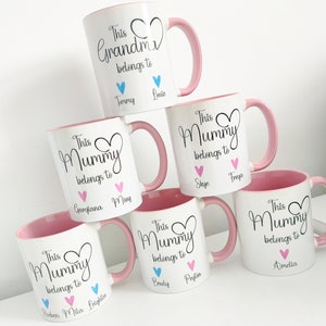 May include: White ceramic mugs with pink handles and interiors. The mugs are personalized with the phrases "This Grandma belongs to" and "This Mummy belongs to", along with names and heart graphics. A stack of mugs.