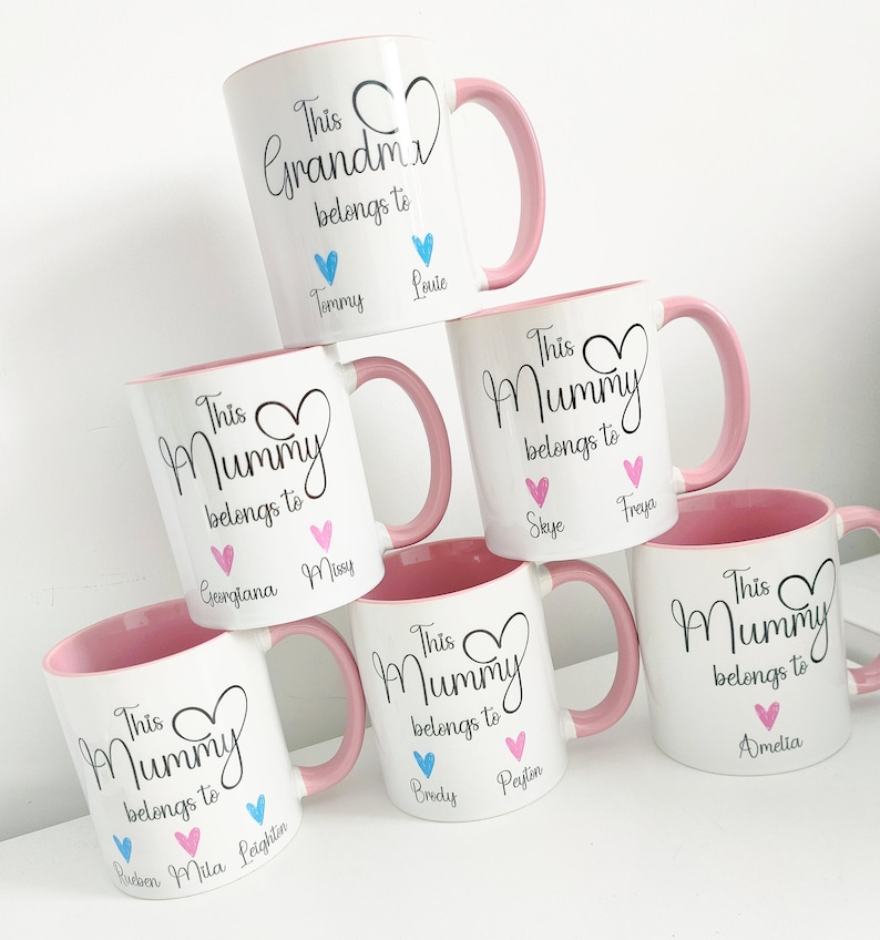 May include: White ceramic mugs with pink handles and interiors. The mugs are personalized with the phrases "This Grandma belongs to" and "This Mummy belongs to", along with names and heart graphics. A stack of mugs.