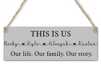 May include: A rectangular gray wooden sign with the text "THIS IS US" in black. Below the text are names and the words "Our life. Our family. Our story." A jute rope is attached for hanging.