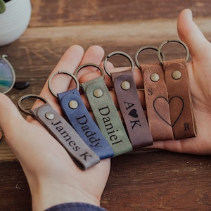 May include: Six leather keychains with different names and designs. The keychains are brown, gray, blue, green, and black. The names on the keychains are James K, Daddy, Daniel, A K, S, and R. One keychain has a heart design.