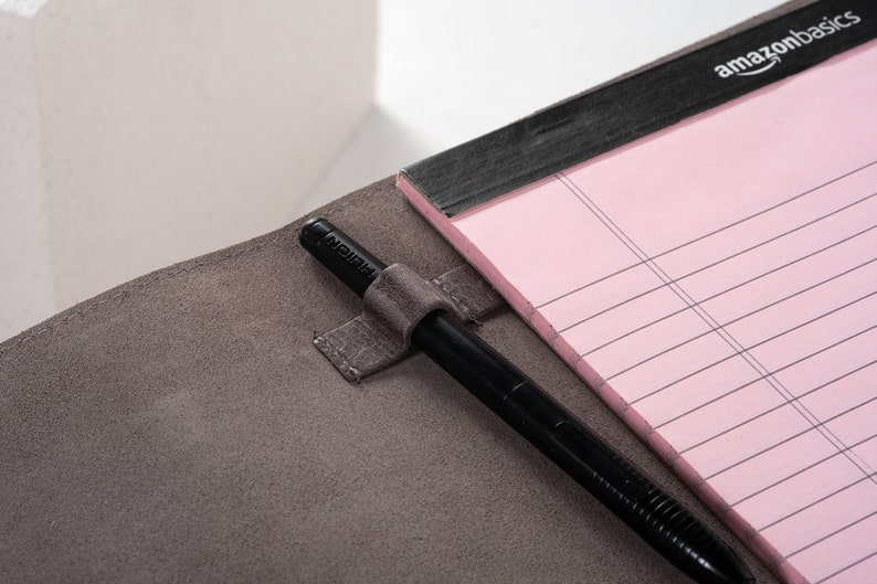 May include: A gray leather notebook with a black pen holder and a pink lined notepad. The notepad has the text "amazonbasics" printed on the cover.