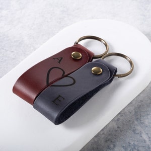 May include: Two leather keyrings with metal rings. The keyring on the left is burgundy with the letter "A" and a heart. The keyring on the right is dark blue with the letter "E" and a heart.
