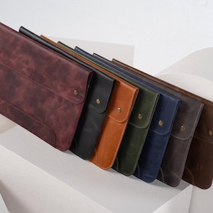 May include: A collection of leather laptop sleeves in various colors, including burgundy, black, brown, and navy. Each sleeve features a flap closure with a brass snap and the embossed word "MARISA". The sleeves are arranged diagonally.