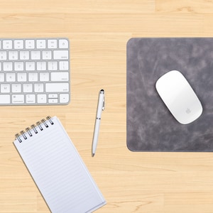 May include: A gray leather mousepad with a white wireless mouse on top. A white keyboard, a white pen, and a spiral bound notebook are also on the wooden desk.