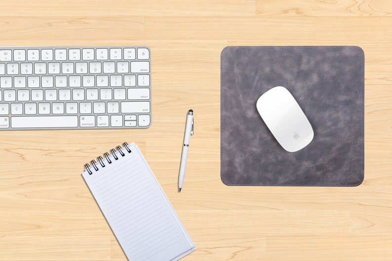 May include: A gray leather mousepad with a white wireless mouse on top. A white keyboard, a white pen, and a spiral bound notebook are also on the wooden desk.