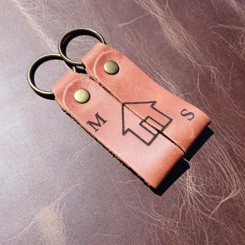 May include: A brown leather keychain with two key rings and a house design in the center. The keychain is engraved with the letters "M" and "S".