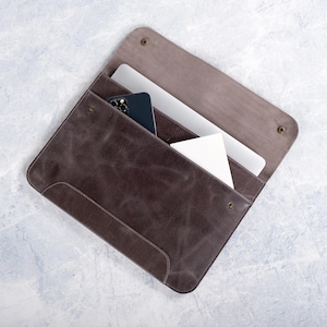 May include: A brown leather laptop sleeve with a zipper closure. The sleeve is open and a laptop and a smartphone are inside.