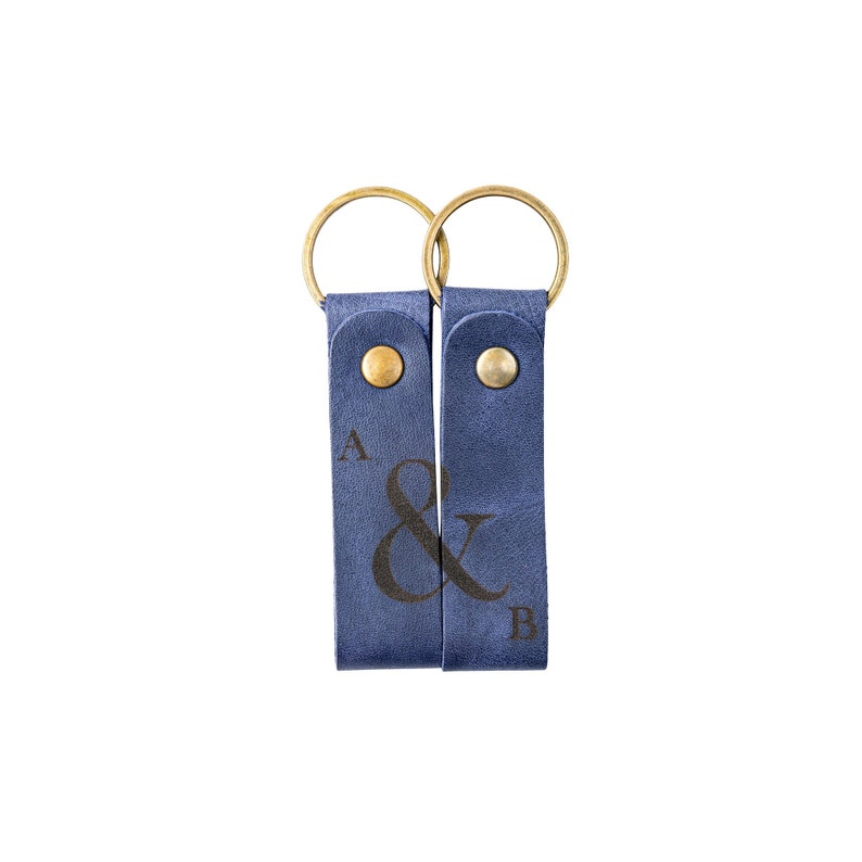 May include: Two blue leather keychains with gold rings and brass snaps. The keychains are connected by a black ampersand symbol and have the letters "A" and "B" engraved on them.