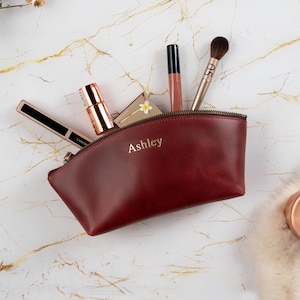May include: A deep red leather cosmetic pouch with the name Ashley embossed in gold lettering on the front. The pouch is open and contains various makeup items including a lipstick, a lip gloss, a makeup brush, and a small compact case.