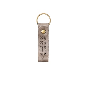 May include: A gray leather keychain with a brass key ring and a heart symbol. The keychain is engraved with the coordinates "31.26.128"N 10.29.17.7"W".