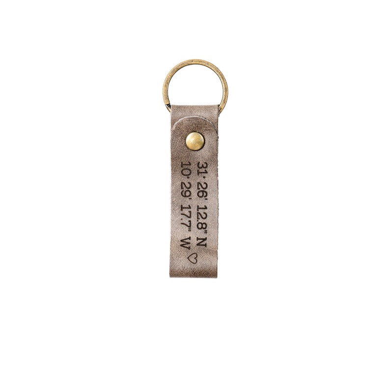 May include: A gray leather keychain with a brass key ring and a heart symbol. The keychain is engraved with the coordinates "31.26.128"N 10.29.17.7"W".