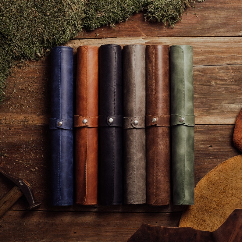 May include: Six rolled-up leather gaming mats in different colors: blue, brown, black, gray, brown, and green. The mats are laid out on a wooden surface.