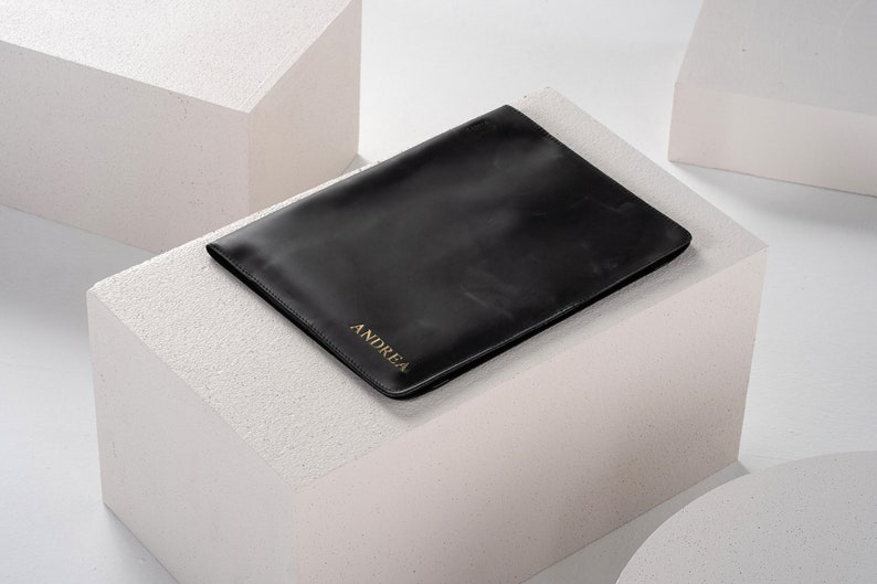 May include: A black leather laptop sleeve with the name "ANDREA" embossed in gold lettering.