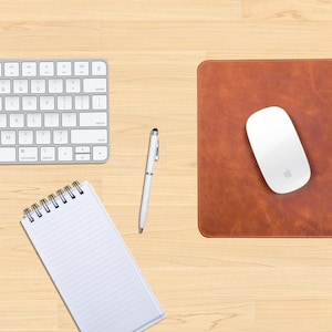 May include: A brown leather desk mat with stitched edges and the initials "AHE" embossed in black. A white wireless mouse is on the mat. A white keyboard and a white pen are on a wooden desk surface.