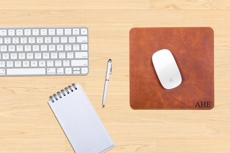 May include: A brown leather desk mat with stitched edges and the initials "AHE" embossed in black. A white wireless mouse is on the mat. A white keyboard and a white pen are on a wooden desk surface.