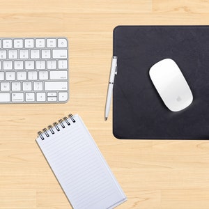 May include: A white wireless keyboard, a black leather mouse pad with a white wireless mouse, a silver pen, and a white spiral bound notebook on a light wood surface.