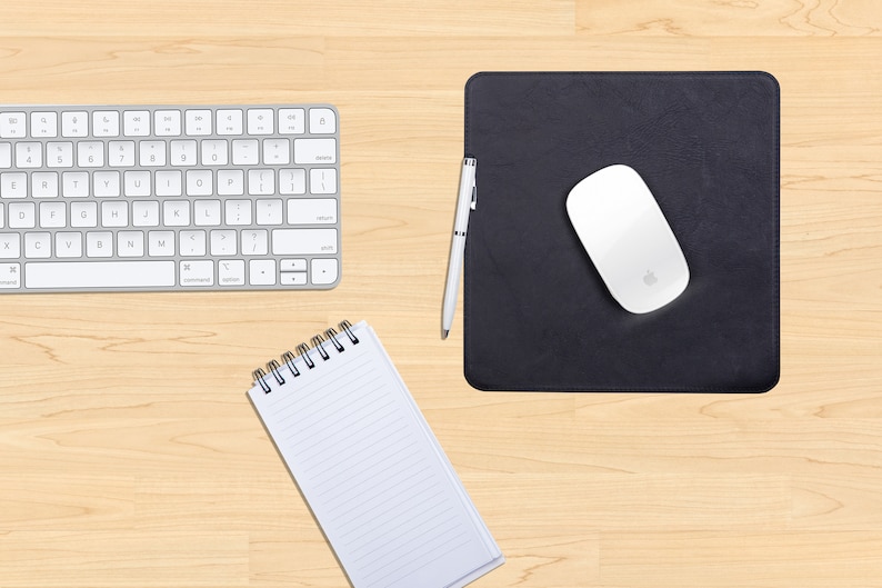 May include: A white wireless keyboard, a black leather mouse pad with a white wireless mouse, a silver pen, and a white spiral bound notebook on a light wood surface.