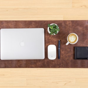 May include: A brown leather desk mat with a laptop, a white mouse, a pen, a small potted plant, a cup of coffee, and a black wallet. The mat has the word "NOVA" printed on it.