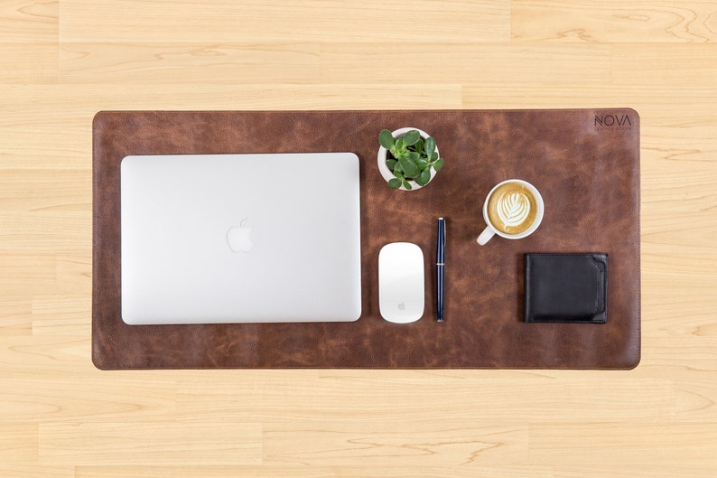 May include: A brown leather desk mat with a laptop, a white mouse, a pen, a small potted plant, a cup of coffee, and a black wallet. The mat has the word "NOVA" printed on it.
