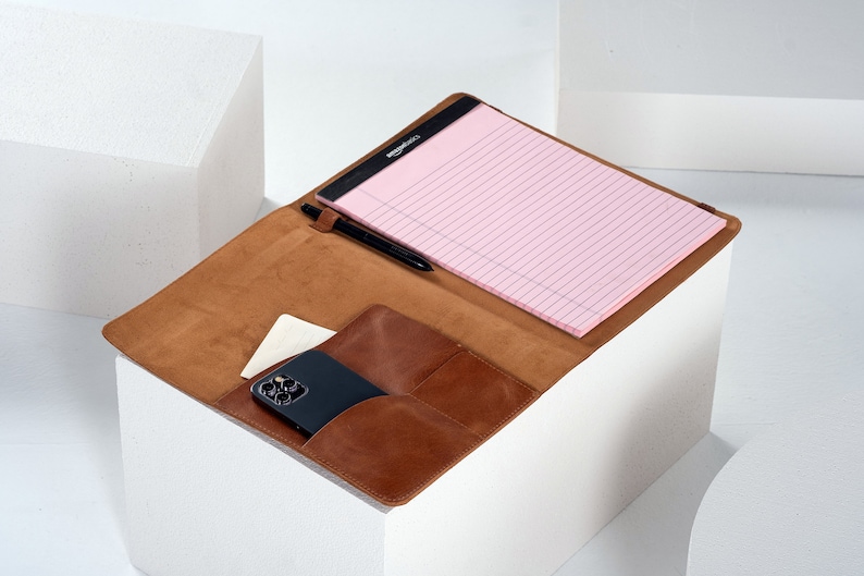 May include: A brown leather portfolio with a notepad, pen, and a smartphone inside. The portfolio has a pocket for a phone and a flap that closes with a snap.