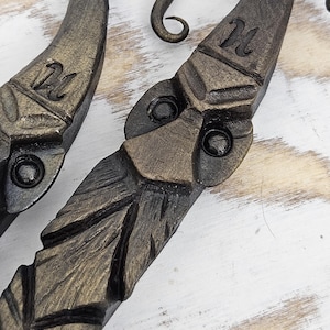 May include: Two black metal hooks with a gold finish. Each hook is shaped like a face with a beard and a small loop at the top. The hooks are marked with the letter "M".