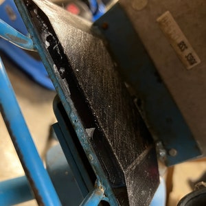 May include: Close-up of a black, textured, triangular object with a metal frame. The object is attached to a light blue metal structure with visible rust. The background is blurred, showing other parts of the structure.