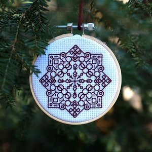 May include: A white embroidery hoop with a brown geometric snowflake design hanging from a branch of a fir tree.