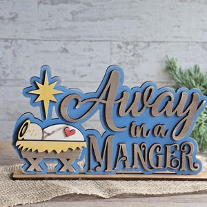 May include: A layered wooden Christmas decoration with the text "Away in a Manger". The design features a star, a baby in a manger, and the words in a blue and brown color scheme. The decoration is on a wooden base.