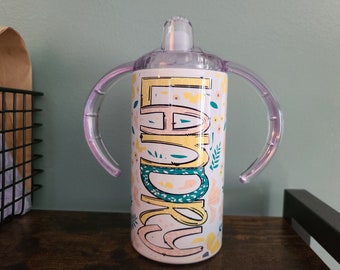 May include: A white stainless steel tumbler with a floral design and the name "Emma" printed on it. The tumbler has two clear plastic handles and a clear plastic lid with a straw.