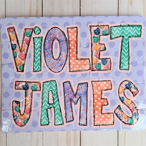 May include: A personalized puzzle with the names "Violet James" in large, colorful letters. The letters are decorated with a pattern of hearts and candy. The puzzle is set against a light purple background with white polka dots.