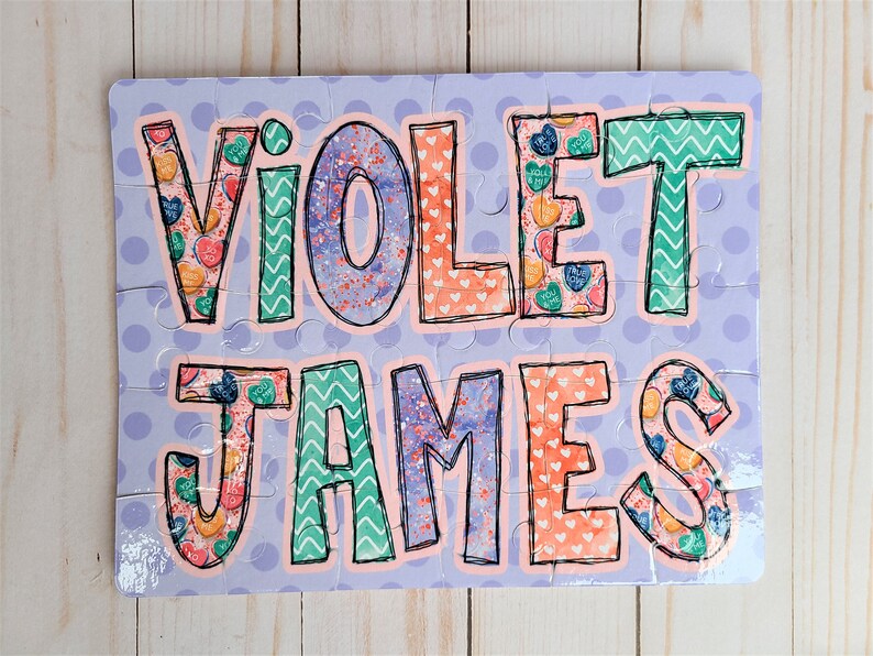 May include: A personalized puzzle with the names "Violet James" in large, colorful letters. The letters are decorated with a pattern of hearts and candy. The puzzle is set against a light purple background with white polka dots.