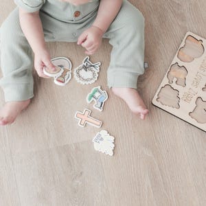 May include: A wooden puzzle with a light brown finish. The puzzle pieces are shaped like a cross, a lamb, a crown of thorns, and other religious symbols. The puzzle is labeled "He Is Risen!" and "God Loves You".