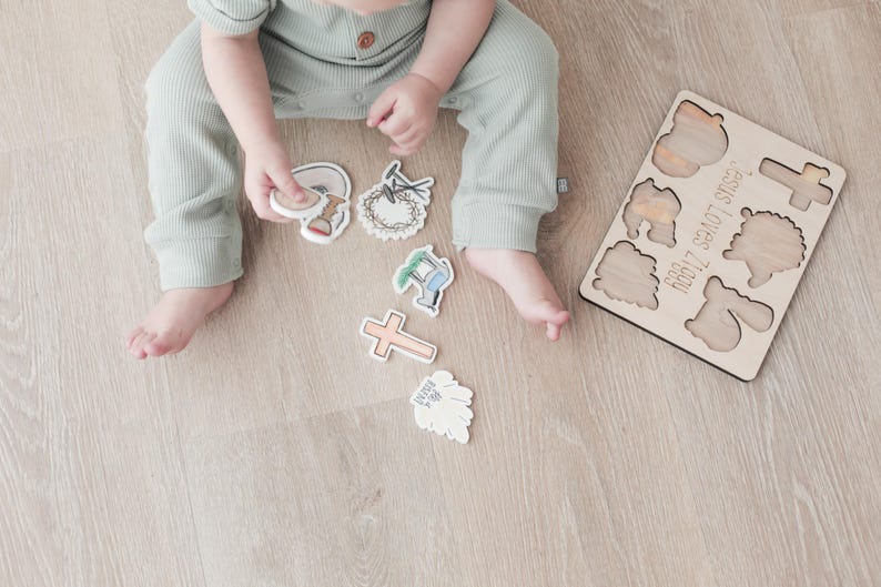 May include: A wooden puzzle with a light brown finish. The puzzle pieces are shaped like a cross, a lamb, a crown of thorns, and other religious symbols. The puzzle is labeled "He Is Risen!" and "God Loves You".