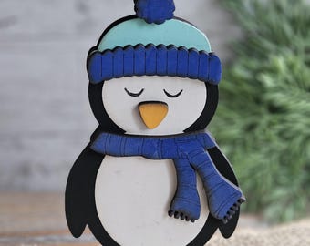 May include: A wooden penguin figurine wearing a blue knit cap and scarf. The penguin has a white belly, black body, and yellow feet. It is standing on a light blue base. The penguin's eyes are closed.