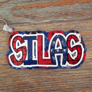 May include: A red, blue, and white keychain with the name "SILAS" printed on it. The keychain features a Spiderman theme with a web pattern and spider silhouettes.