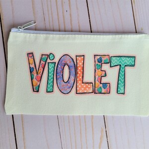 May include: A light yellow canvas pencil case with a zipper closure. The name "VIOLET" is printed on the front in colorful patterned letters.