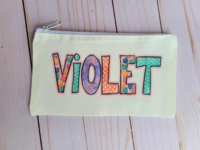 May include: A light yellow canvas pencil case with a zipper closure. The name "VIOLET" is printed on the front in colorful patterned letters.