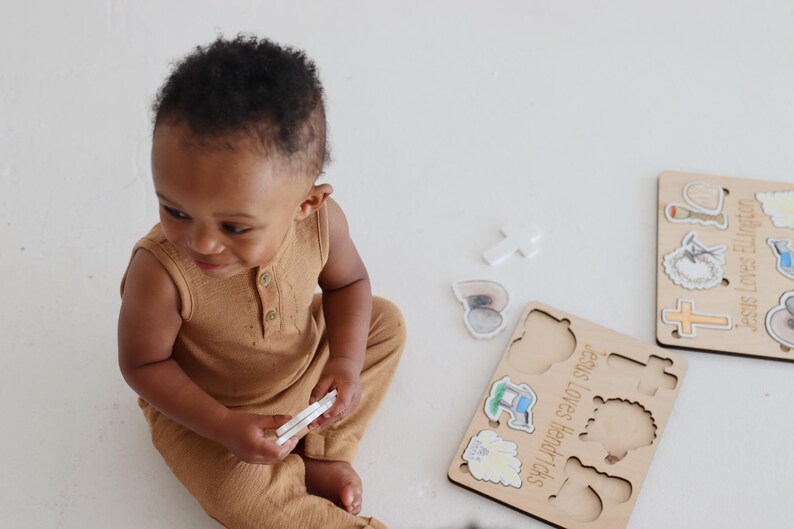 May include: A wooden puzzle with colorful illustrations of religious symbols. The puzzle pieces are shaped like the symbols and fit into the corresponding cutouts on the board. The text on the board reads "Jesus Loves Little Lambs".