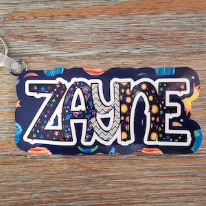 May include: A blue and black key chain with the name "Zayne" written in white letters. The letters are decorated with a space-themed pattern of stars, planets, and rockets.