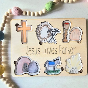 May include: A wooden puzzle with a light brown background and a white border. The puzzle pieces are cut out in the shape of various Christian symbols, including a cross, a crown of thorns, a chalice, a loaf of bread, a tomb, a donkey, and a sunburst. The puzzle also includes the text "Jesus Loves Parker".