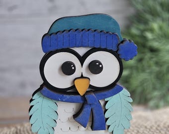 May include: A wooden owl figurine with a blue hat, scarf, and base. The owl has large white eyes with black pupils, a yellow beak, and light blue wing details. The figurine is on a light blue base.