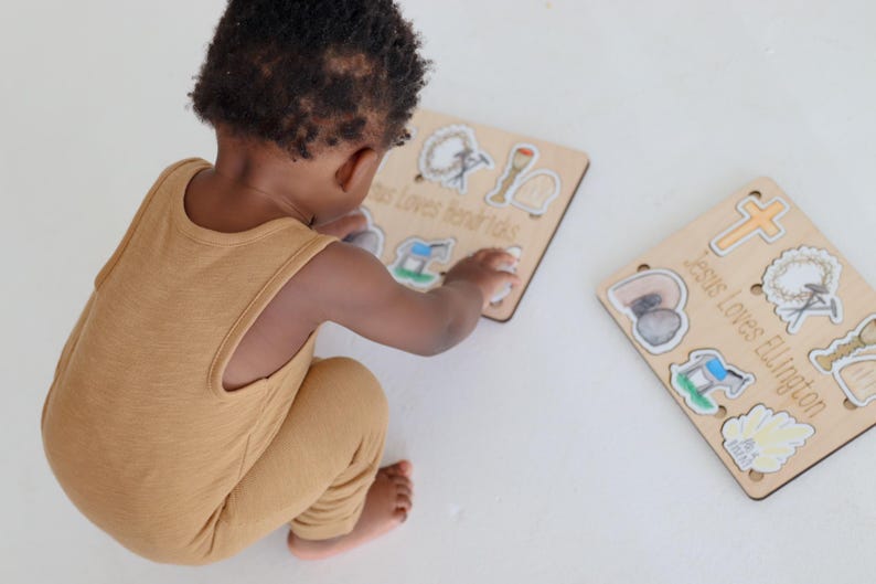 May include: A child plays with a wooden puzzle set featuring images of religious symbols. The puzzle pieces are labeled "Jesus Loves Little Kids" and "Jesus Loves Ellington".